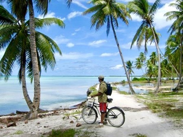Mike Pate riding a bicycle along a sandy track on a palm-lined Pacific atoll