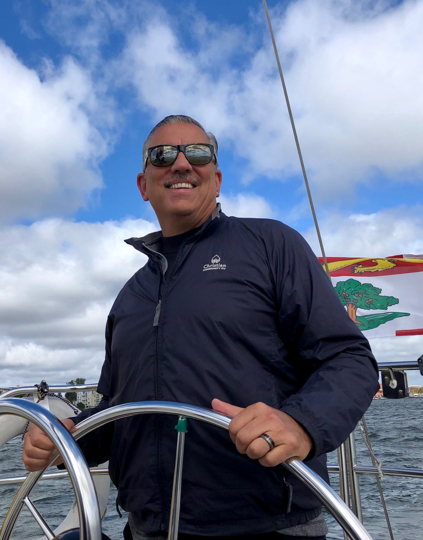 Mike at the wheel of a sailboat in sunglasses, smiling under a sky full of clouds
