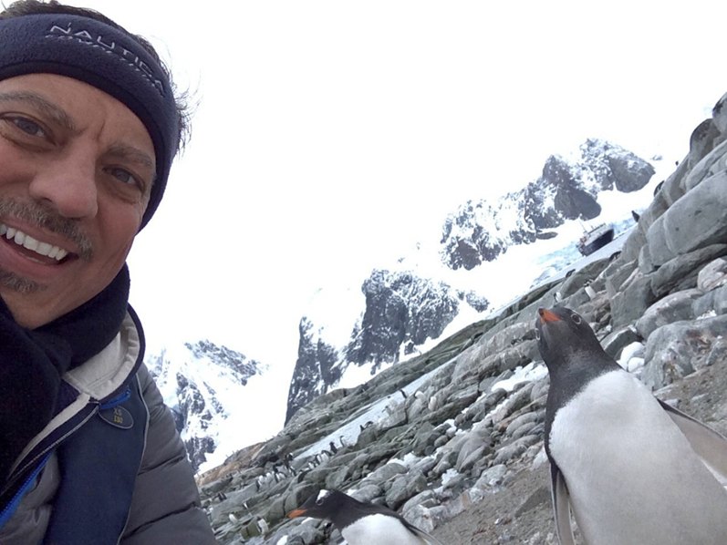 Selfie of Mike in a knit hat with a gentoo penguin standing beside him on a rocky Antarctic shore